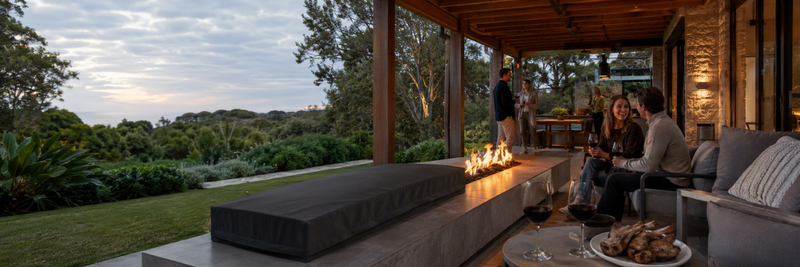 Wide outdoor entertaining area with guests, wine and food beside a long gas fire table under a timber covered patio overlooking a garden.