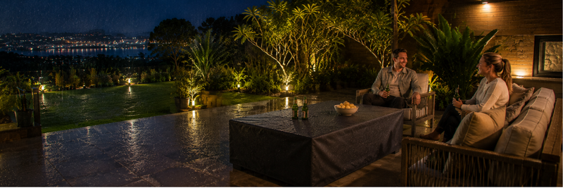 Rain falls across a modern Australian alfresco patio at night while a rectangular gas fire table sits protected beneath a fitted black weatherproof cover, with a couple relaxing nearby overlooking a lit garden and distant water views.