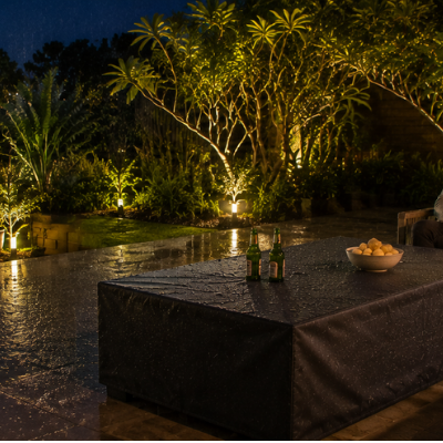 Rain falls across a modern Australian alfresco patio at night while a rectangular gas fire table sits protected beneath a fitted black weatherproof cover, with a couple relaxing nearby overlooking a lit garden and distant water views.