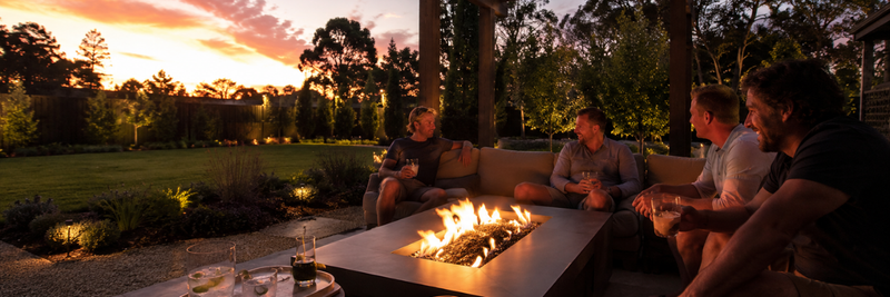 A group of men relaxing around a large gas fire table beneath a covered pergola at sunset, overlooking a landscaped backyard with outdoor lighting.