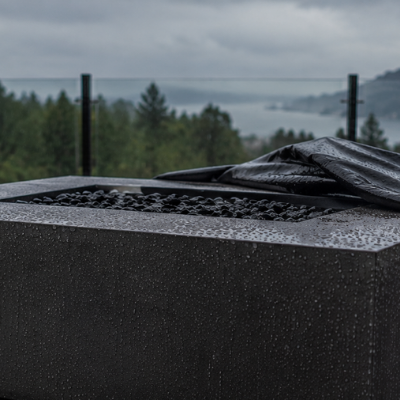 Outdoor propane fire table on a rain-soaked patio with water droplets and a partially fitted protective cover under an overcast sky.