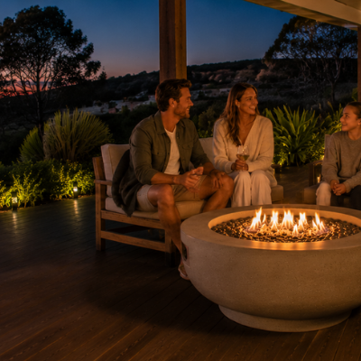 Wide coastal alfresco entertaining area with a family gathered around a round fire bowl at dusk, overlooking bushland views and a glowing Australian sunset.