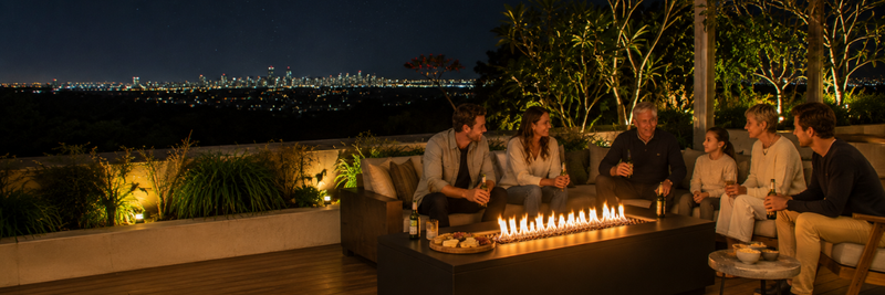 Wide evening view of a family gathered around a modern fire table on a spacious Australian outdoor deck overlooking city lights, with warm landscaping, snacks, and relaxed conversation creating a welcoming atmosphere.