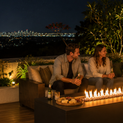 Wide evening view of a family gathered around a modern fire table on a spacious Australian outdoor deck overlooking city lights, with warm landscaping, snacks, and relaxed conversation creating a welcoming atmosphere.