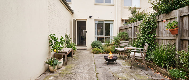 A small fire pit burning in a narrow paved townhouse courtyard beside a timber fence