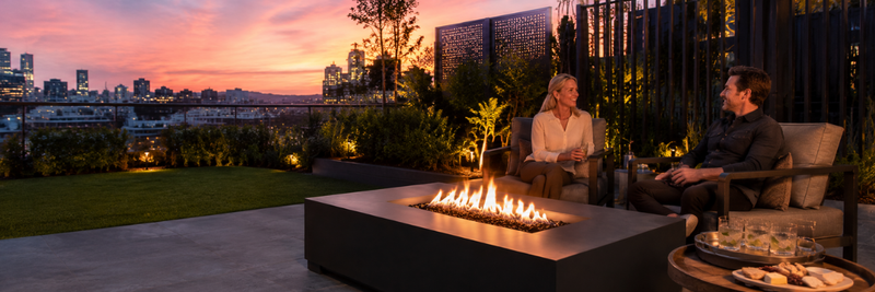 A couple relaxing beside a low rectangular gas fire table on a modern rooftop patio at sunset, with city skyline views and outdoor lounge seating.