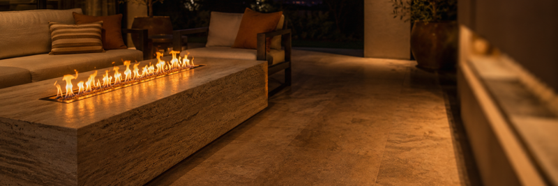 A premium travertine ethanol fire table burns with a long amber flame on a sheltered alfresco terrace at dusk, casting warm light across stone pavers and outdoor seating.