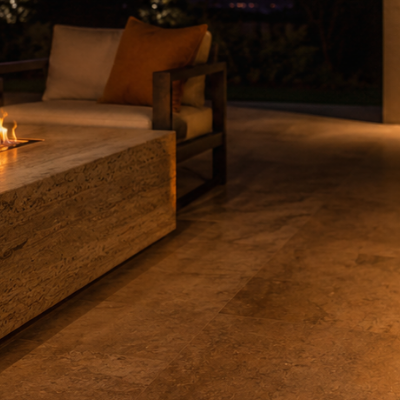 A premium travertine ethanol fire table burns with a long amber flame on a sheltered alfresco terrace at dusk, casting warm light across stone pavers and outdoor seating.