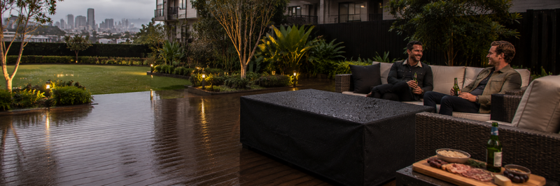 Wide view of a covered rectangular fire table on a wet timber deck in a modern Australian backyard while two men relax with drinks beside outdoor lounges and a grazing platter during light rain.