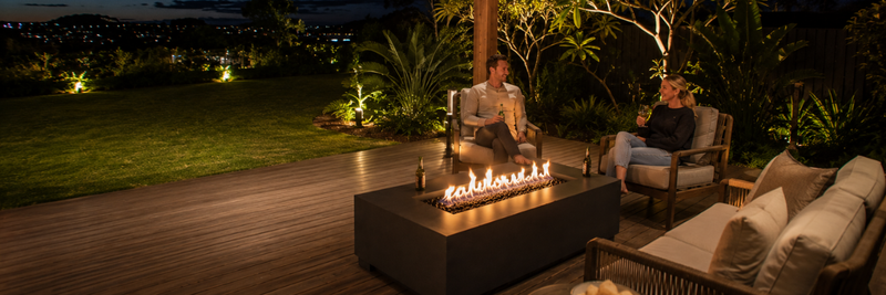 Wide nighttime view of a modern outdoor entertaining area with a rectangular gas fire table positioned safely between lounge seating and open deck space, surrounded by illuminated gardens and lawn.