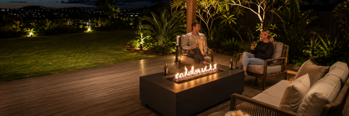 Wide nighttime view of a modern outdoor entertaining area with a rectangular gas fire table positioned safely between lounge seating and open deck space, surrounded by illuminated gardens and lawn.