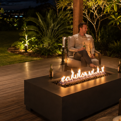 Wide nighttime view of a modern outdoor entertaining area with a rectangular gas fire table positioned safely between lounge seating and open deck space, surrounded by illuminated gardens and lawn.