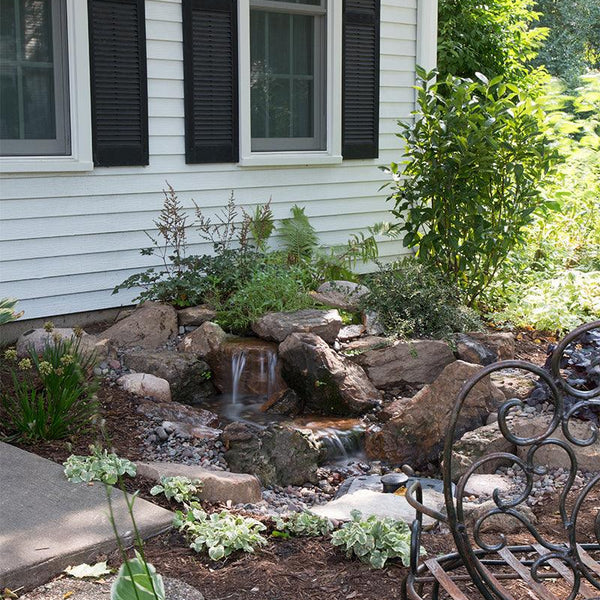 A small pondless waterfall built with the Aquascape DIY Backyard Waterfall Kit, cascading over rock into gravel beside a house
