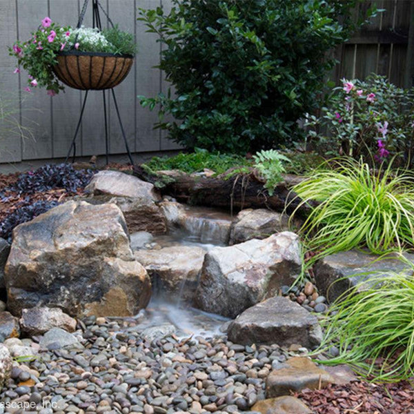 A backyard waterfall fountain built with the Aquascape Landscape Fountain Kit, water disappearing into a pebble bed surrounded by planting