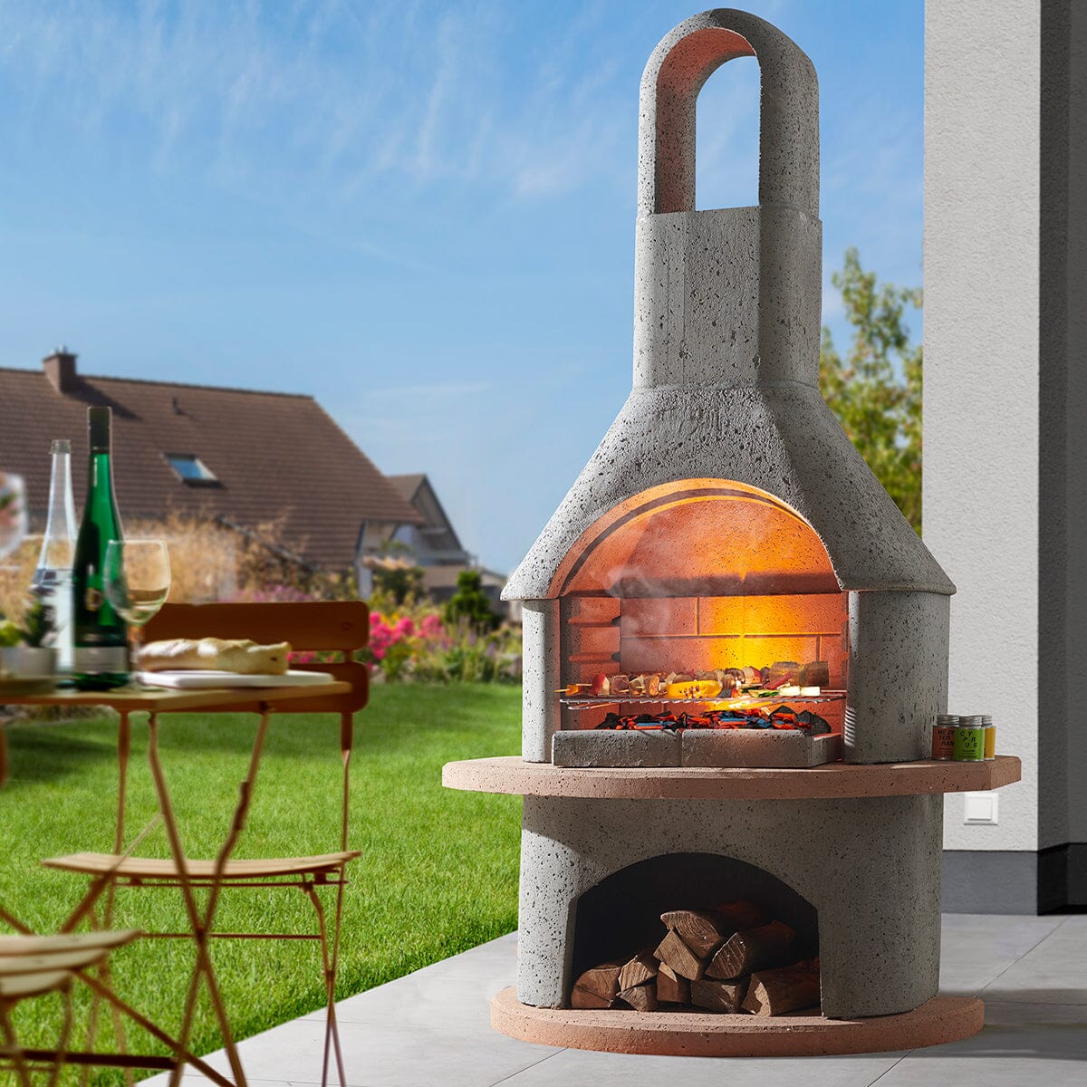 Buschbeck Carmen lit during the day on a patio, with the fire glowing under the arch and logs stored below