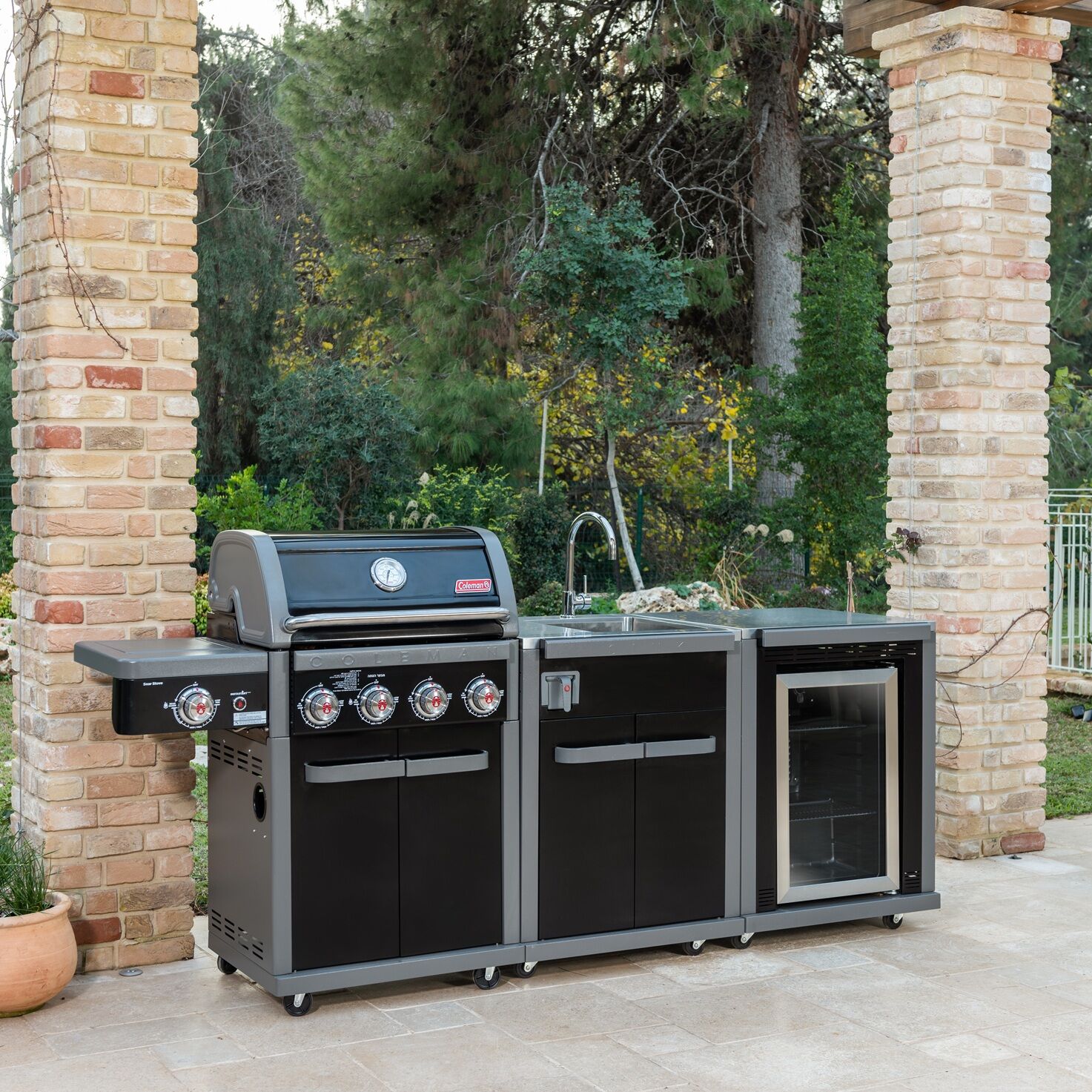 Coleman Revolution 4 Burner outdoor kitchen with sink and fridge modules beside a brick pillar