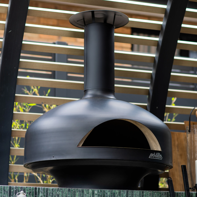 Black Polito Giotto Oven BTS installed in a modern outdoor kitchen beneath timber battens with curved chimney and wood fired cooking chamber visible