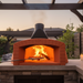 A close-up of a built-in Valoriani Family TOP120 Pizza Oven in an outdoor kitchen. A fresh pizza is being slid into the oven using a metal peel. Vibrant flames from the wood fire lick the top of the internal dome, illustrating the professional cooking performance of this Valoriani pizza oven Australia model.