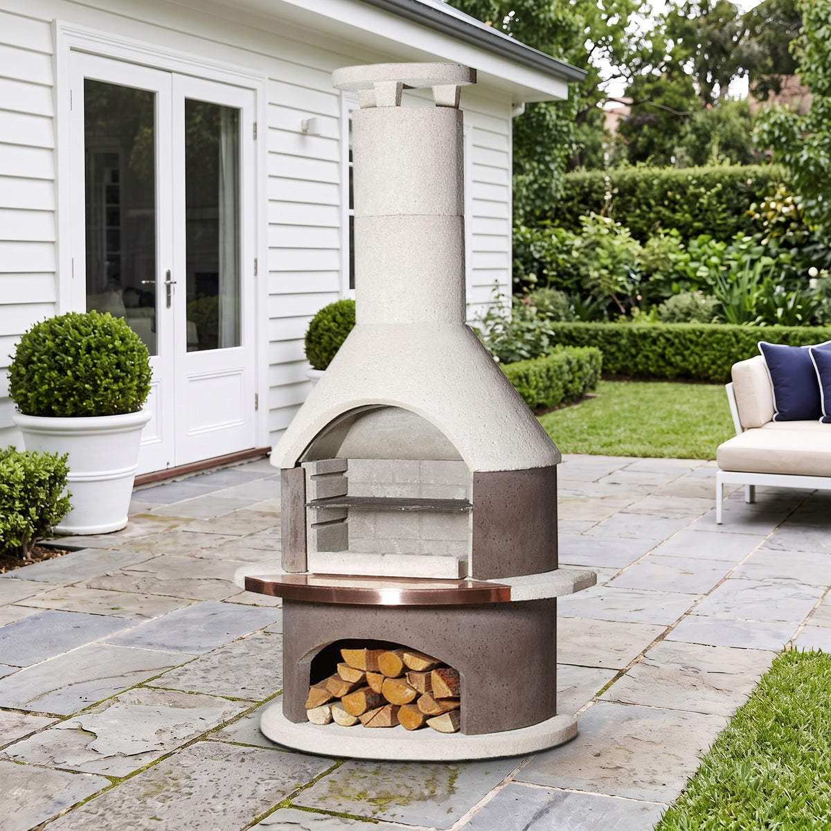 Buschbeck Rondo 3-in-1 outdoor fireplace, BBQ and pizza oven in Mocha with copper shelf insert, on a bluestone terrace beside a white weatherboard home