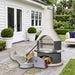 Buschbeck Rondo 3-in-1 outdoor fireplace, BBQ and pizza oven kit in Grey with pizza oven door, base extension, peels and cutter, on a bluestone terrace beside a white weatherboard home