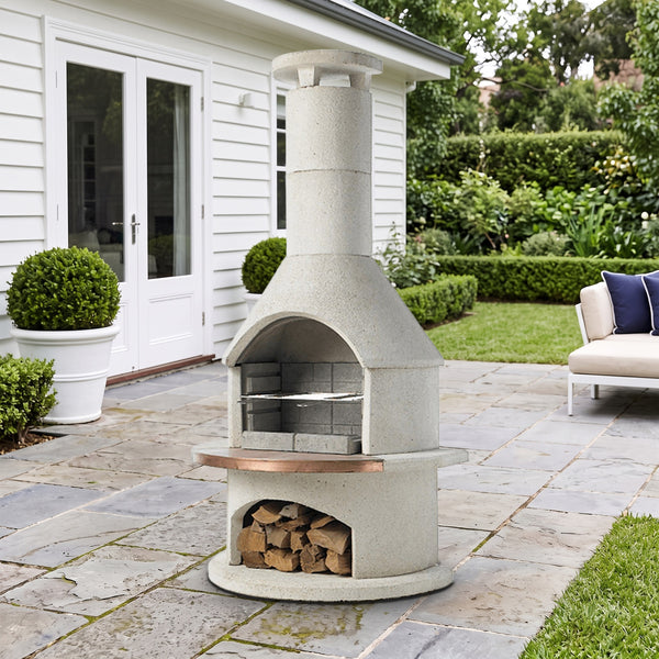 Buschbeck Rondo 3-in-1 outdoor fireplace, BBQ and pizza oven in White with copper shelf insert, on a bluestone terrace beside a white weatherboard home