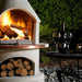 Buschbeck St Moritz Outdoor Fireplace lit at night beside a table set with wine and a cheese board