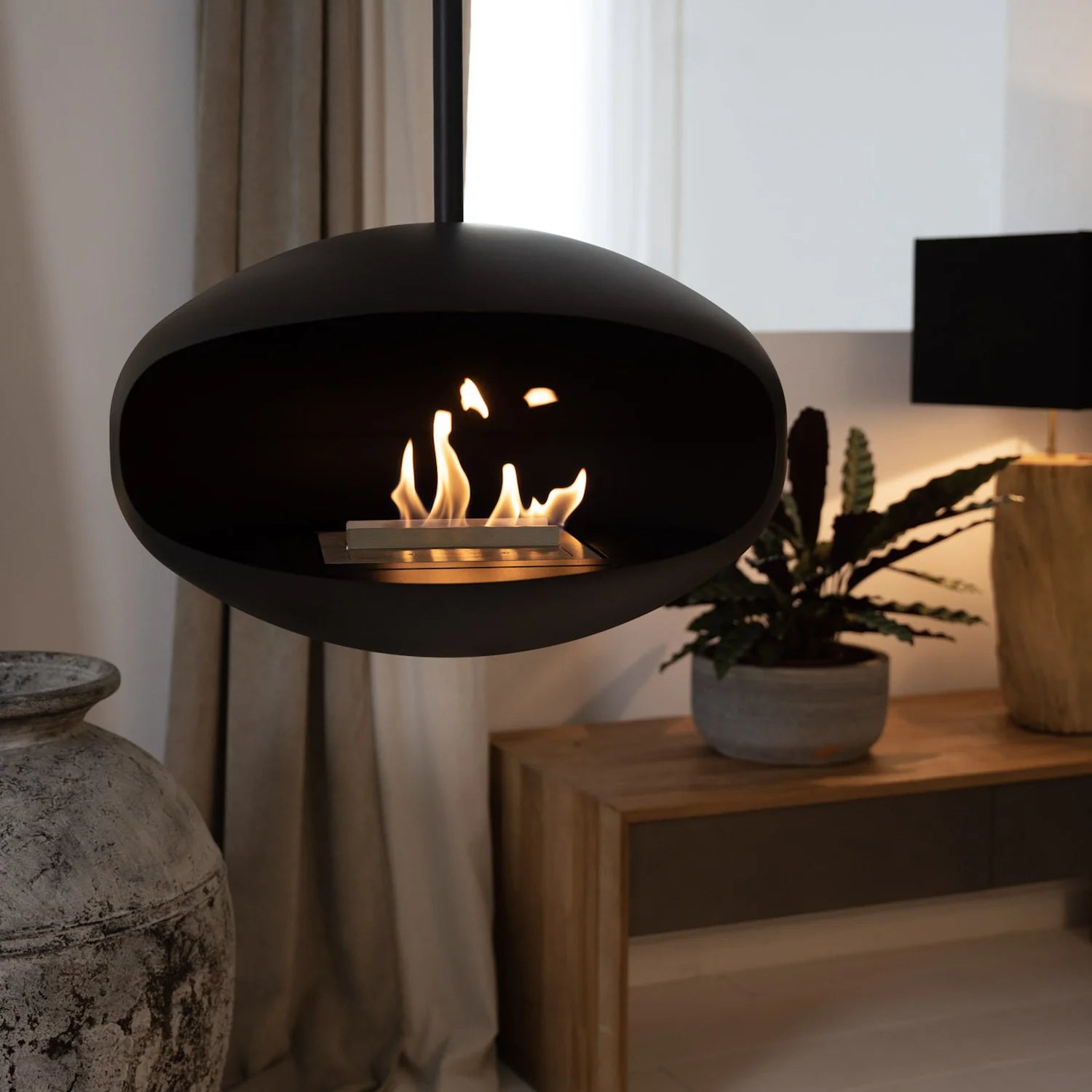 Cocoon Aeris Hanging Bioethanol Fireplace in matt black hanging in a living room beside a timber console