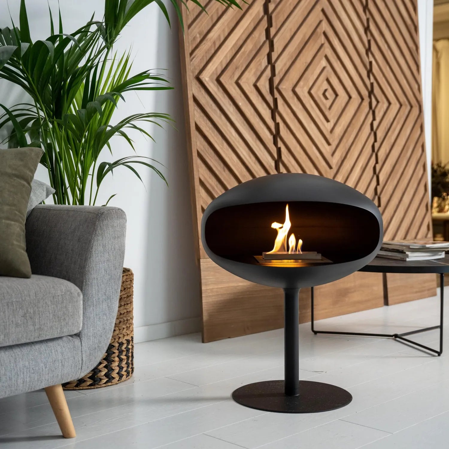 Cocoon Pedestal Bioethanol Fireplace in matt black beside a grey armchair