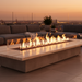 EcoSmart Linear 130 Fire Pit Kit placed on a sleek rooftop patio with a clear view of the city skyline at sunset, flames flickering from the fire pit centerpiece.