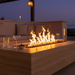 EcoSmart Linear 50 Fire Pit Kit with Black Burner in a stylish outdoor setting during dusk, flames illuminating the space, complemented by cocktails and a relaxing lounge area, showcasing the fire pit's design and ambiance.