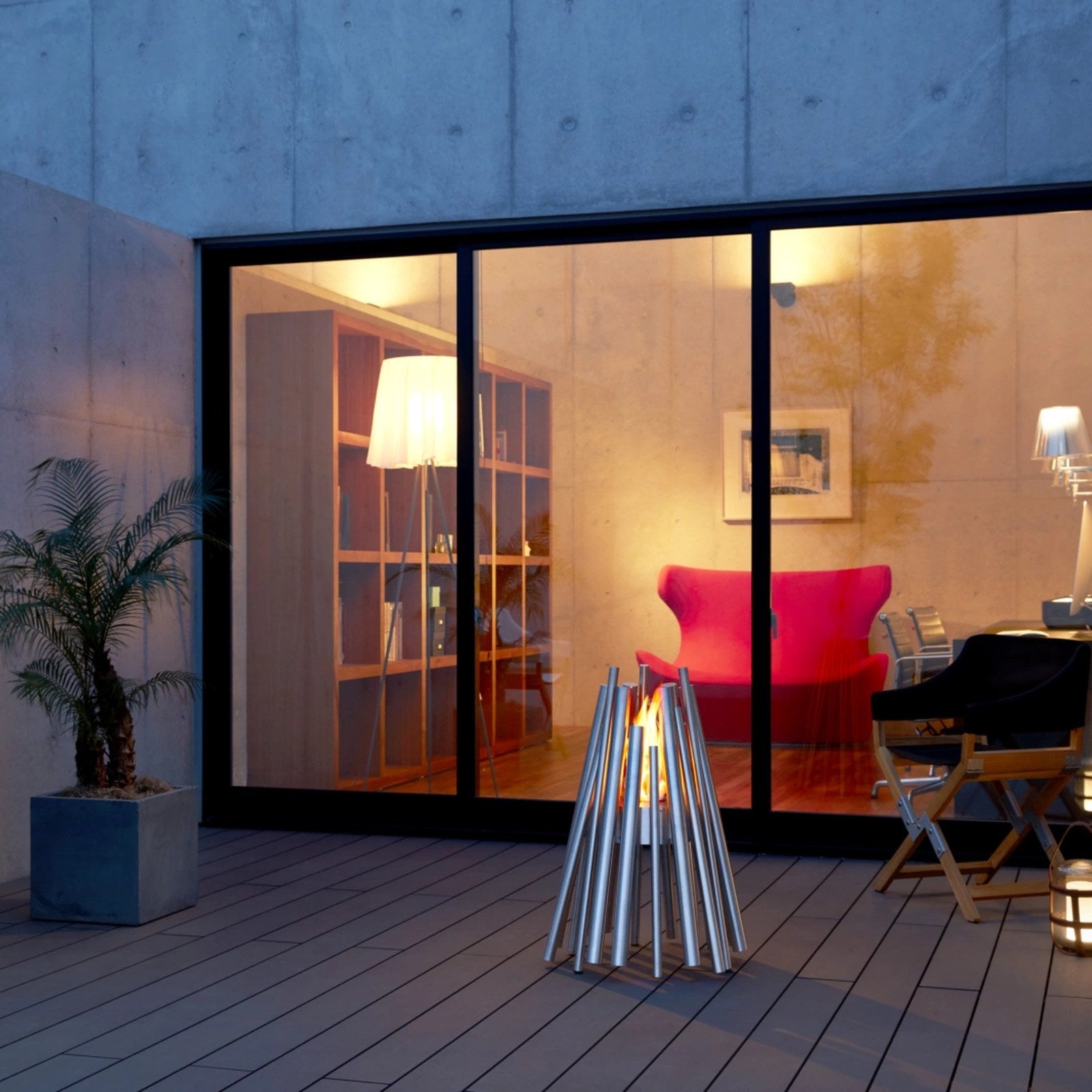 EcoSmart Stix fire pit lit in a concrete courtyard at dusk beside a designer armchair.