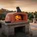 An external pizza oven setup featuring the Valoriani Family TOP120 Pizza Oven wood-burning model. The oven is built onto a rustic stone pedestal in a lush garden during sunset. Bright orange flames from burning wood logs fill the interior, and a wooden pizza paddle rests nearby on the stone patio.