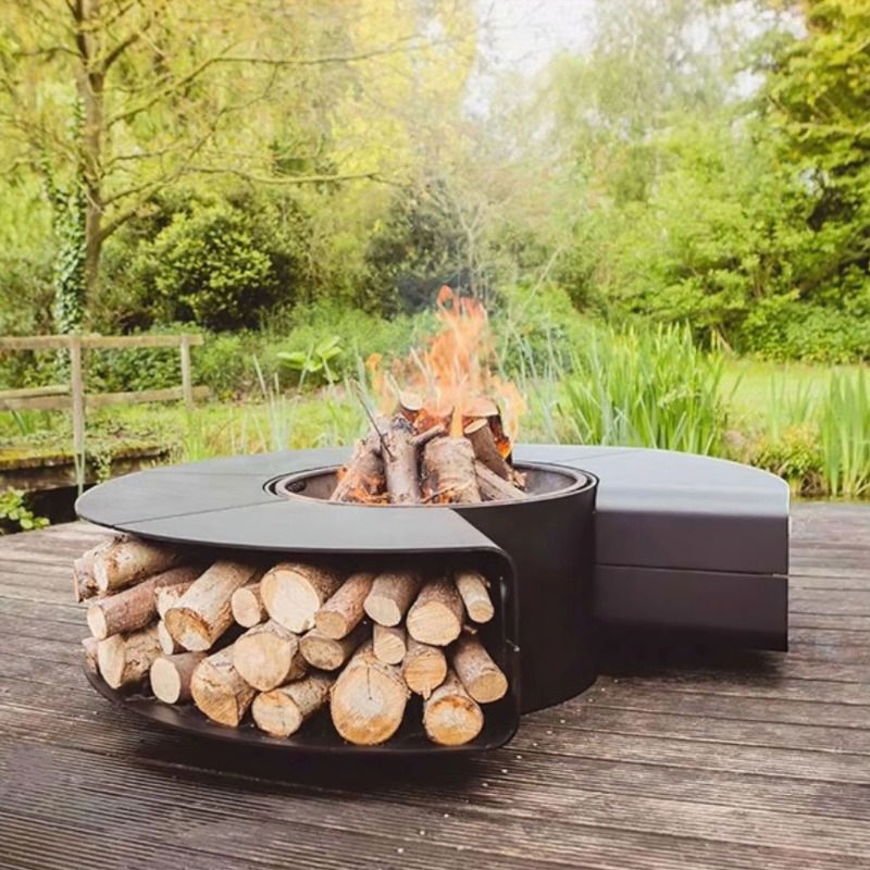 Fabrizio Fire Pit on a wooden deck in a natural garden setting, with stacked logs below and a live fire in the centre, styled for outdoor relaxation and entertaining.