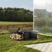 Low-profile Fabrizio Fire Pit styled as a sculptural outdoor fire pit table, shown on a lawn beside a reflective wall with integrated firewood storage and tabletop surface.