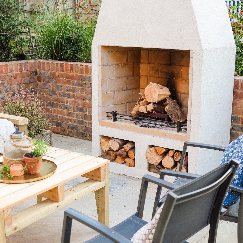 Wide backyard view of the Ochre Living Hotham Complete Fireplace Kit in an outdoor entertaining space with pergola lights, lounge seating, coffee table, and garden beds, showcasing outdoor fireplace ideas Australia.