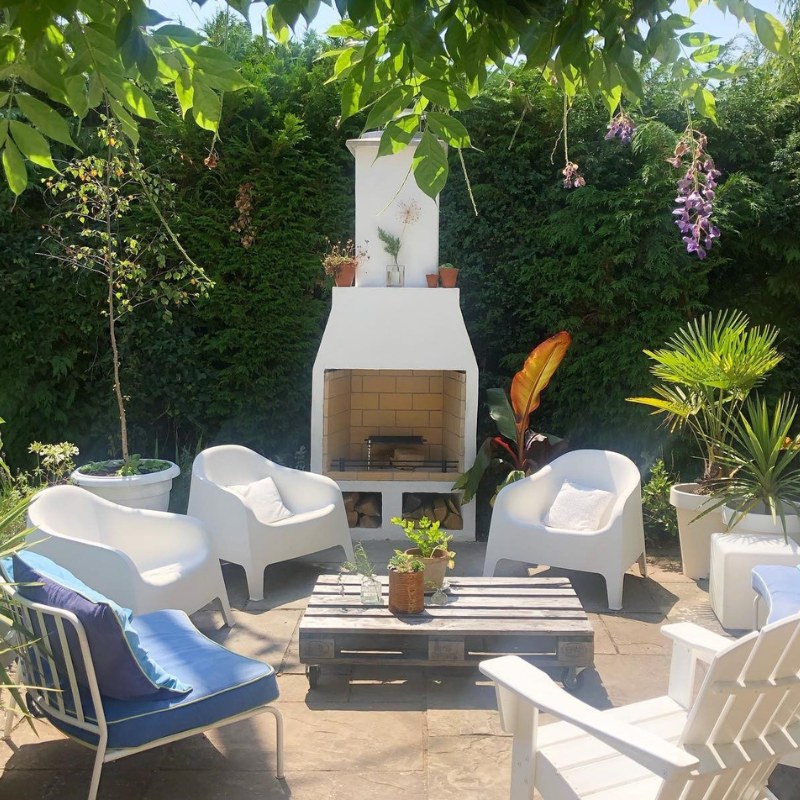 Dark-finish version of the Ochre Living Hotham Complete Fireplace Kit installed on a timber deck with pergola posts, slatted privacy screen, and black outdoor furniture in a modern forest setting.