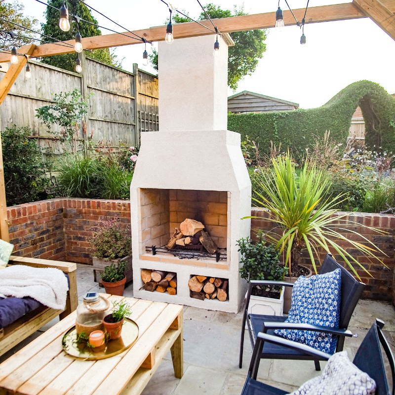 Ochre Living Hotham Complete Fireplace Kit with a large fire burning in a backyard patio setting with pergola lighting, lounge furniture, and stacked logs below, showing outdoor fireplace kits Australia in use.