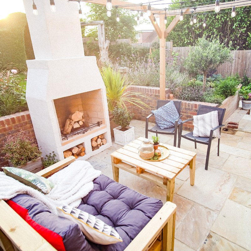 Front view of the Ochre Living Hotham Complete Fireplace Kit installed in a garden patio under string lights with surrounding plants, chairs, and wood storage, showing an outdoor fireplace with chimney Australia.