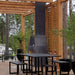 Stone-clad version of the Ochre Living Hotham Complete Fireplace Kit installed in a covered brick patio with benches, fireplace tools, and stacked wood, showing premium outdoor fireplace ideas Australia.