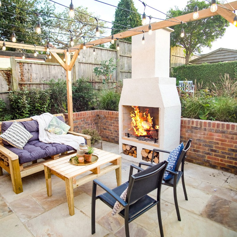 Styled garden courtyard featuring the Ochre Living Hotham Complete Fireplace Kit surrounded by white outdoor chairs, potted plants, and a low table, presenting relaxed outdoor fireplace ideas Australia.