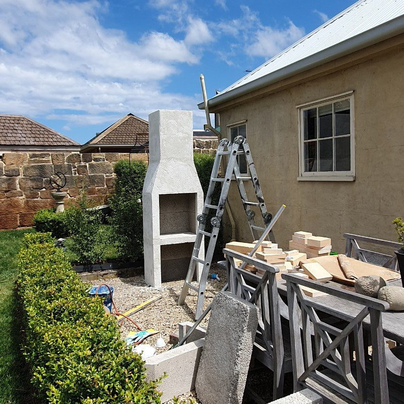 The Ochre Living Ossa Complete Fireplace Kit assembled outdoors before rendering, showing the fireplace structure and chimney during installation in an Australian backyard.