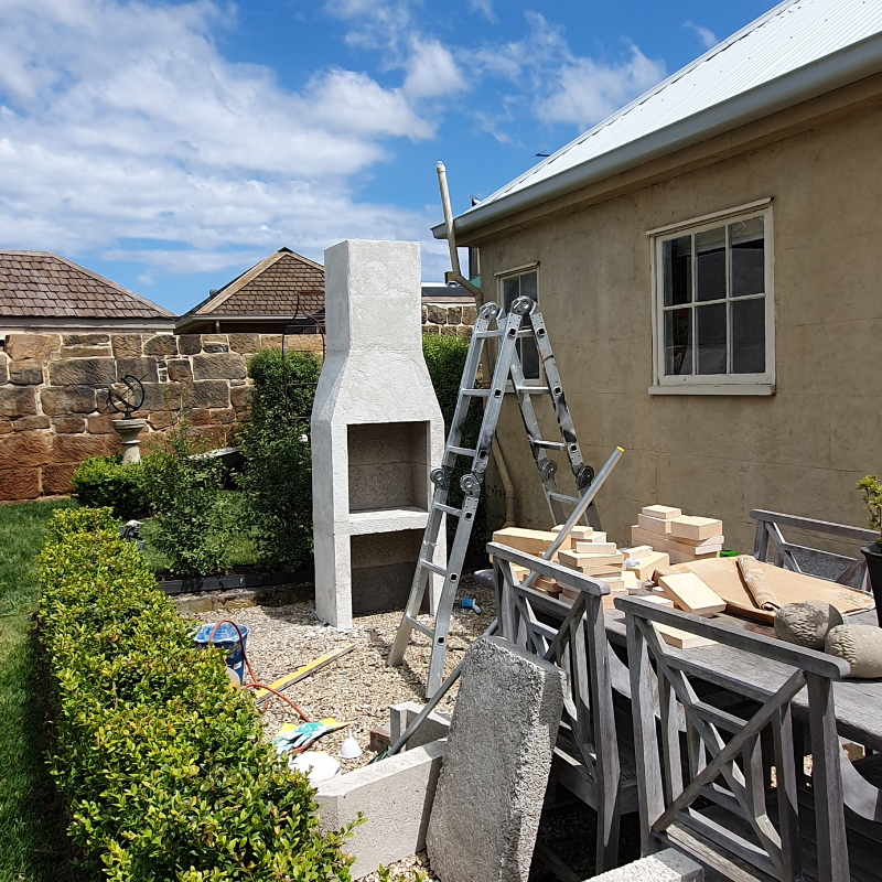 The Ochre Living Ossa Complete Fireplace Kit assembled outdoors before rendering, showing the fireplace structure and chimney during installation in an Australian backyard.