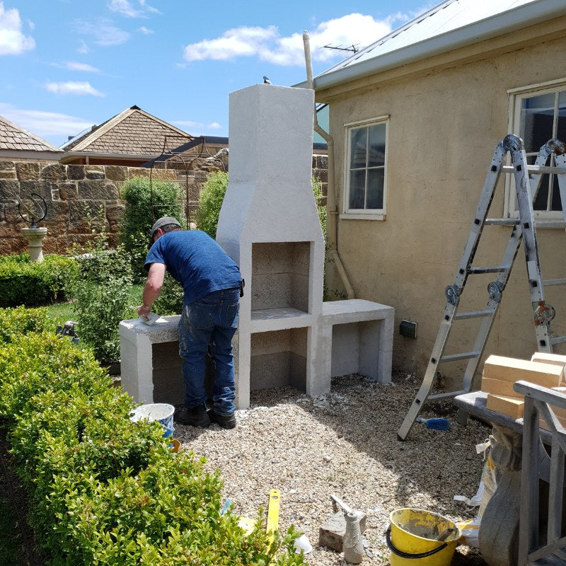 Wide garden view of the Ochre Living Ossa Complete Fireplace Kit during installation and rendering, with tools, ladder, and firebricks visible beside the outdoor fireplace.