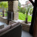 Side view of a finished Ochre Living Ossa Complete Fireplace Kit installed under a covered outdoor entertaining area, showing the chimney, glass side screen, bench seating, and landscaped garden in an Australian backyard.