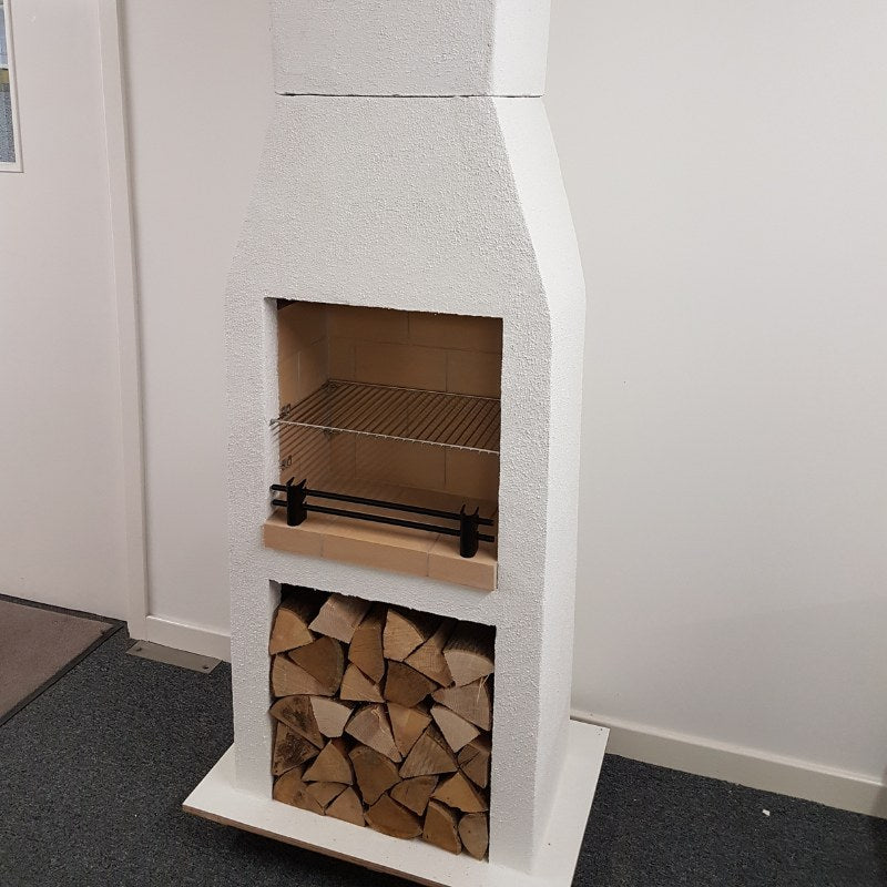 Front view of the Ochre Living Ossa Complete Fireplace Kit displayed indoors in a showroom, with stacked firewood below and a cooking grill fitted inside the firebox.