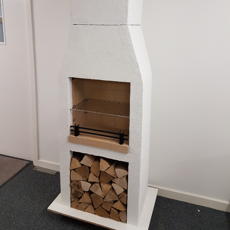 Front view of the Ochre Living Ossa Complete Fireplace Kit displayed indoors in a showroom, with stacked firewood below and a cooking grill fitted inside the firebox.