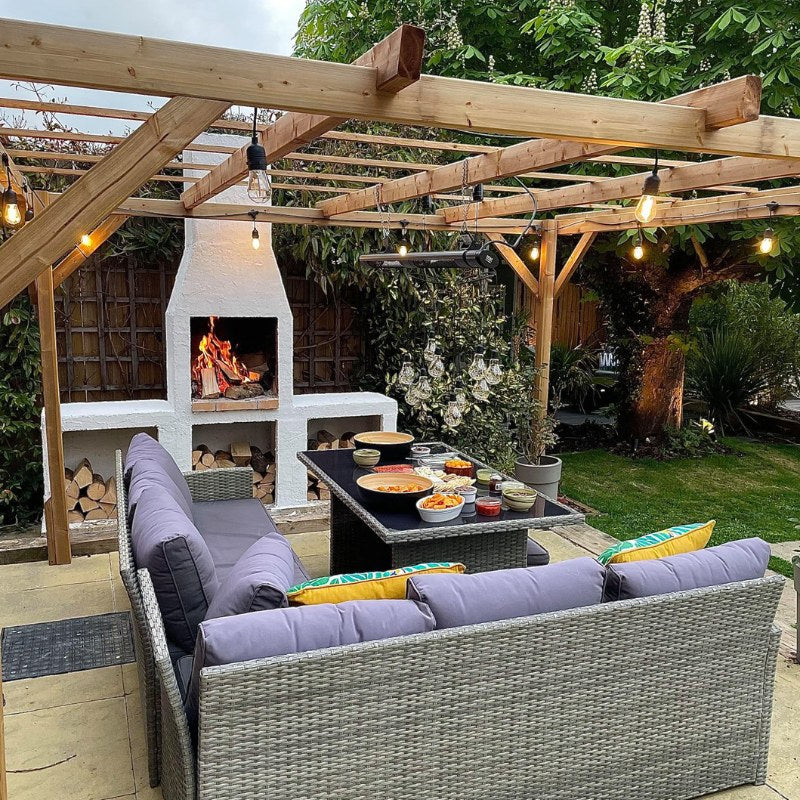 Outdoor entertaining area with a pergola and lighting, featuring the Ochre Living Ossa Complete Fireplace Kit burning firewood, with built-in log storage and seating, demonstrating outdoor fireplace ideas in Australia for backyard dining and entertaining.