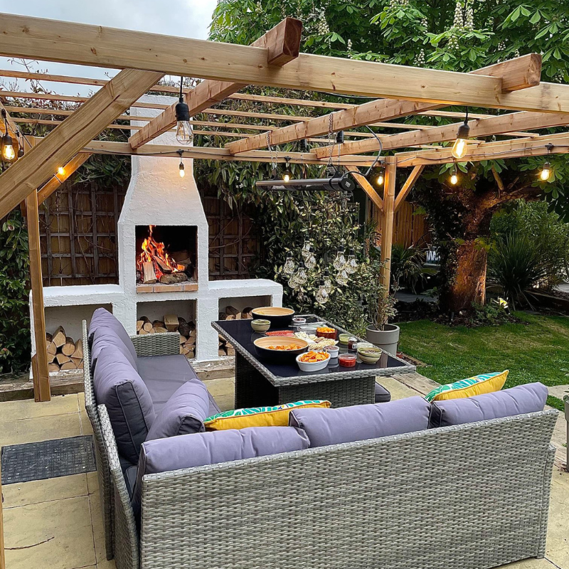 Outdoor entertaining area with a pergola and lighting, featuring the Ochre Living Ossa Complete Fireplace Kit burning firewood, with built-in log storage and seating, demonstrating outdoor fireplace ideas in Australia for backyard dining and entertaining.