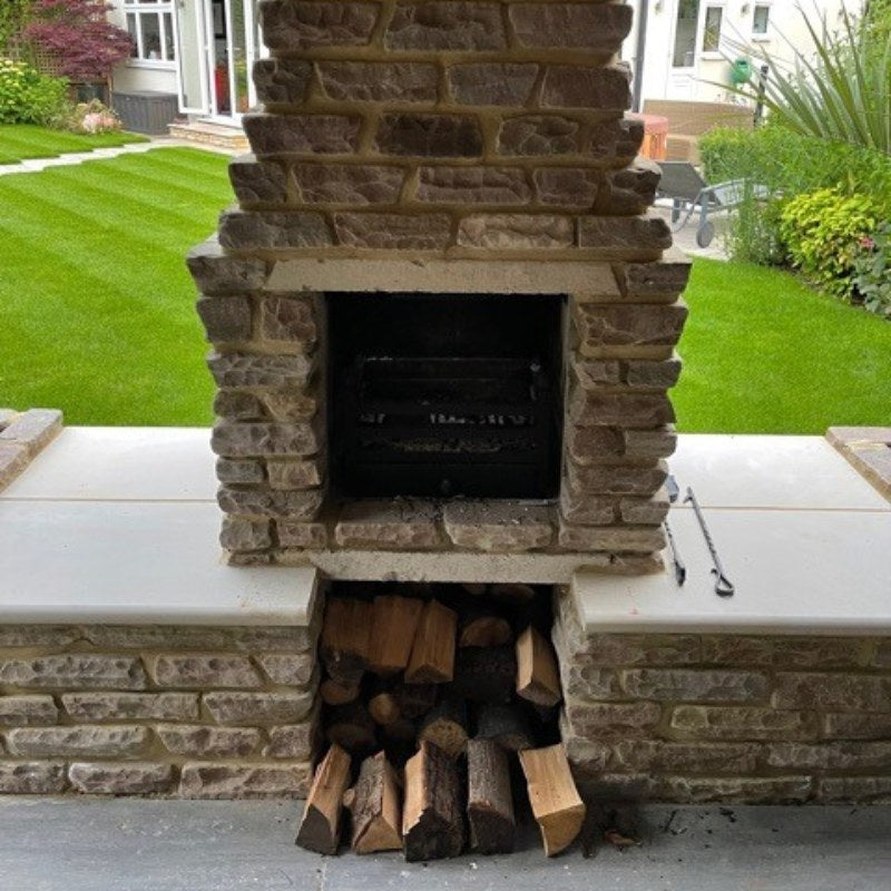Front view of an installed Ochre Living Ossa Complete Fireplace Kit with stone cladding, log storage underneath, and built-in bench surrounds in a landscaped backyard patio setting, suitable for outdoor fireplace ideas in Australia.