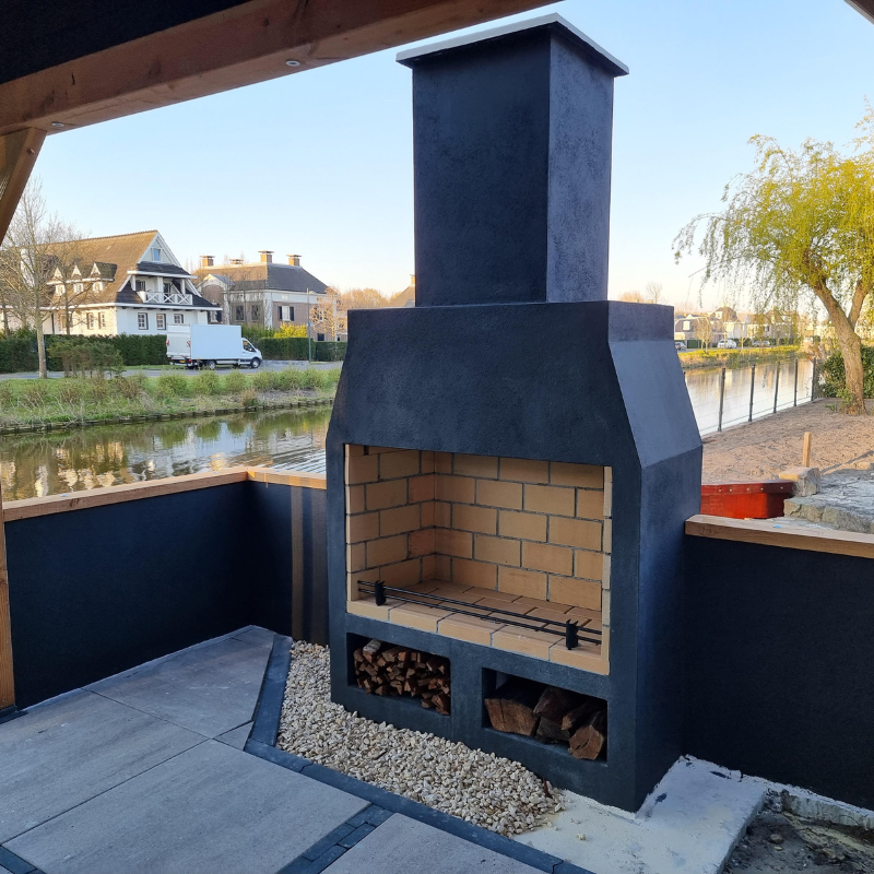 Ochre Living Perisher Complete Fireplace Kit in a dark rendered finish, installed on a covered outdoor area beside a canal with wood storage beneath the firebox.